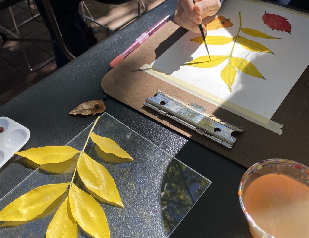 Photograph of a person's hands as they paint leaves with watercolors in golden hues. Next to them are real leaves as a subject