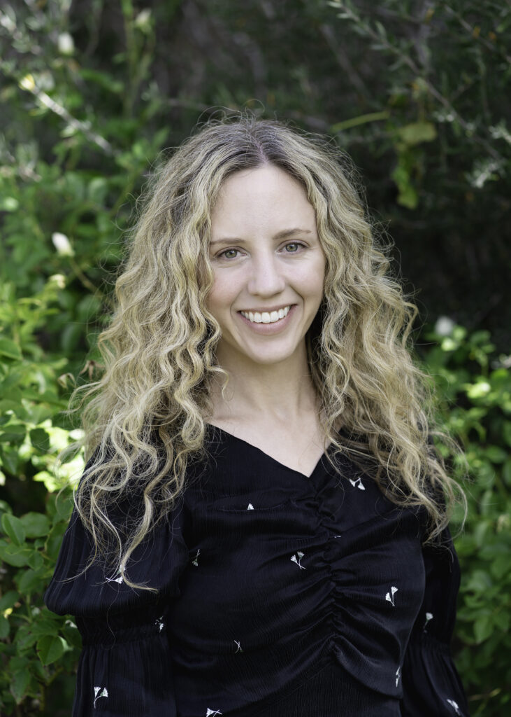 Portrait of a person with long curly light hair smiling and looking at the camera. They wear a dark shirt and behind them are green foliage.