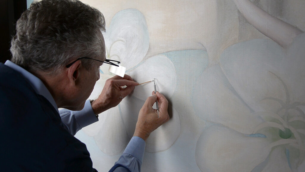 Photograph of a person with a special eye glass looking closely at a large painting of flowers by O'Keeffe and using small tools to repair it.