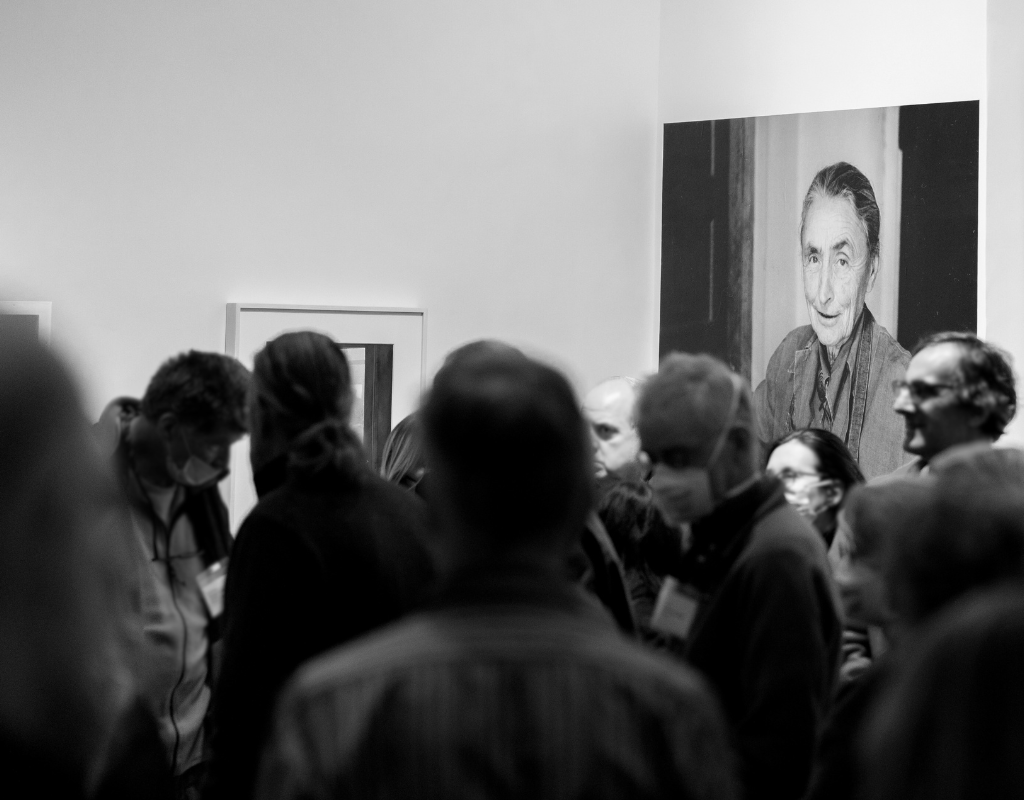 Black and white photograph of a crowd in the foreground and blurry and a photograph of O'Keeffe in the background and in focus.