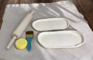 Photograph of two small white ceramic serving dishes handmade. On the left of the dishes are a brush, sponge, and rolling pin.