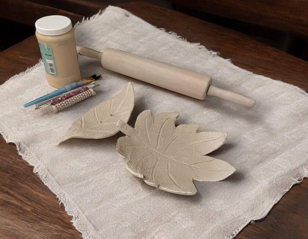 Photograph of two small flat bowls made of clay in the shape of leaves. They rest on a cloth on a wooden table and nearby are small tools