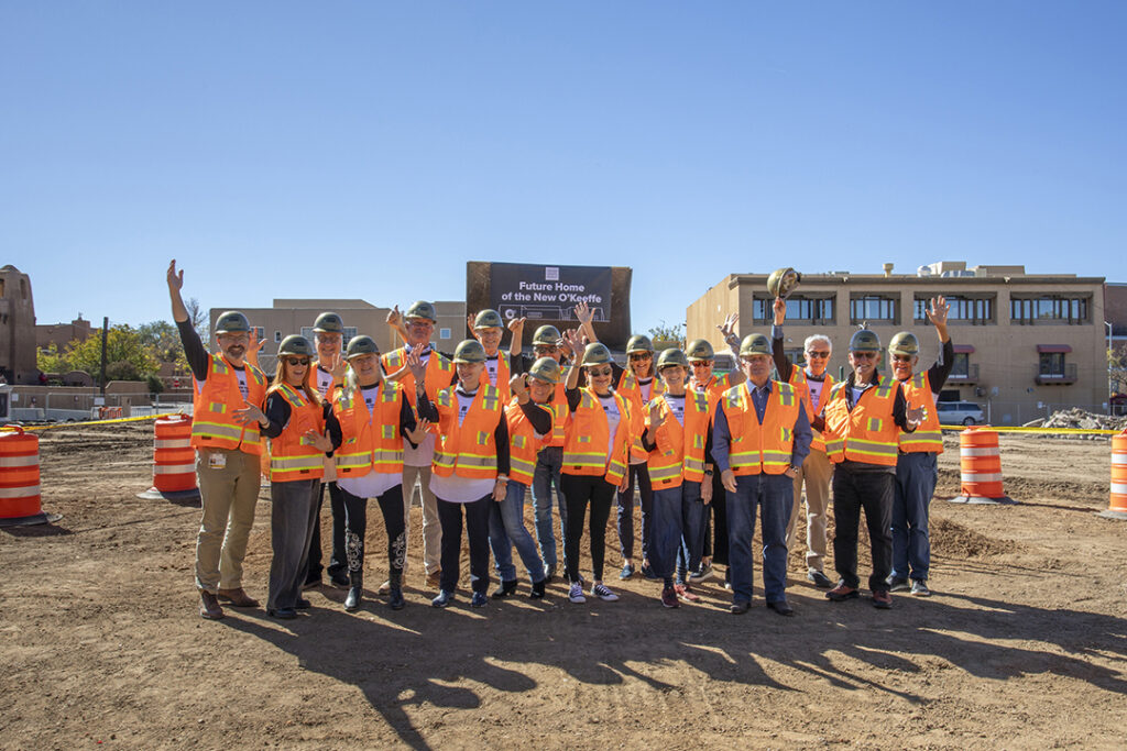 Group of people in construction hats and bright orange safety bests standing on a construction site. Behind them is a large banner with the words "Future home of the new O'Keeffe." A blue sky above.  