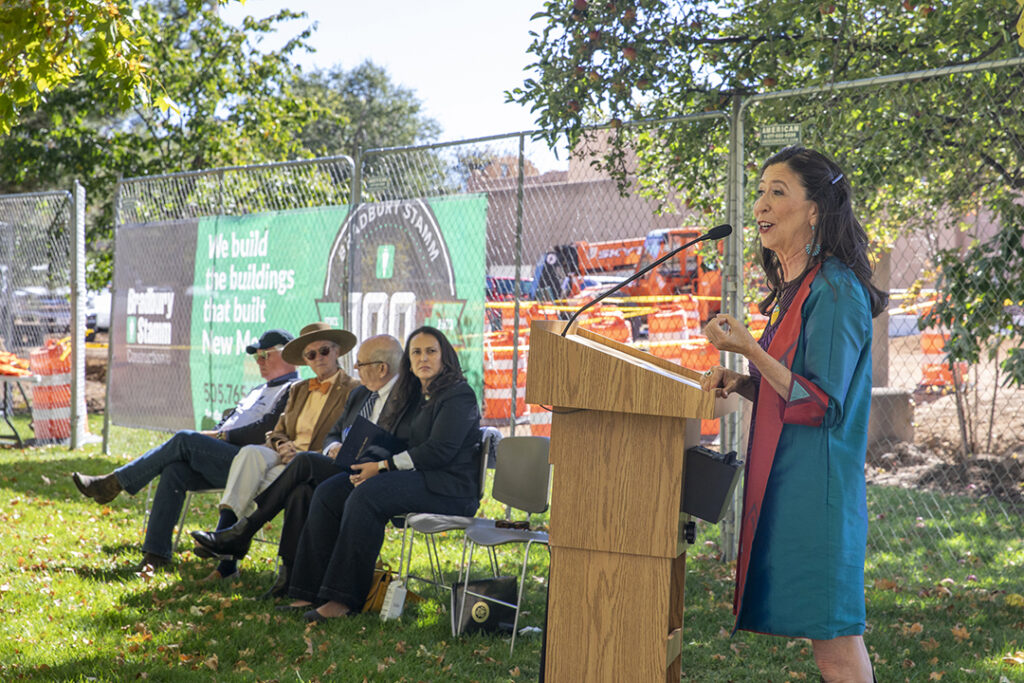 US Congresswoman Teresa Leger Fernandez standing at a podium speaking in front of a construction site. A few people sit nearby on a green lawn. 