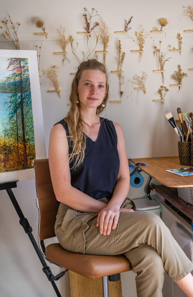 Photograph of a person with long light hair sitting with their hands crossed a table in an artist's studio. Behind them on the wall are several different types of dried plants pinned up for reference.