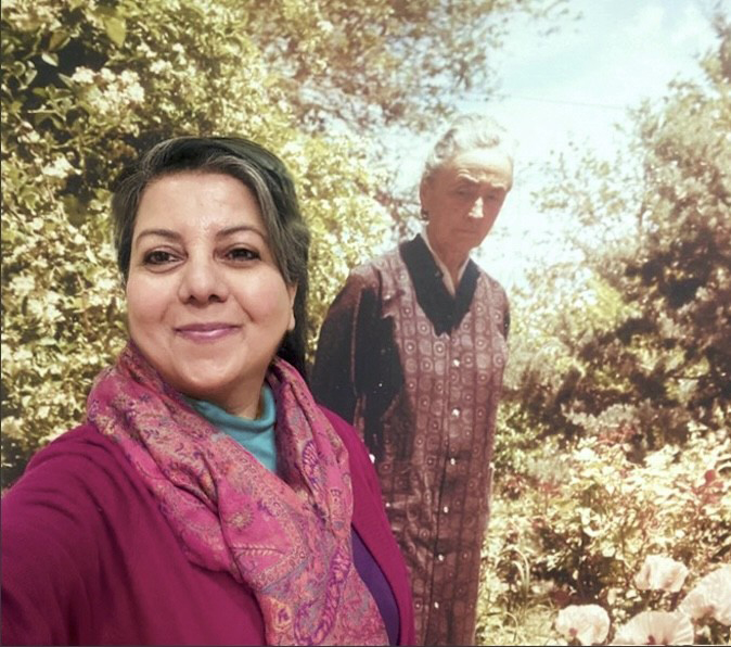 Photograph of a person with dark hair tied up smiling in front of a large photograph of Georgia O'Keeffe. The person wears a colorful scarf.