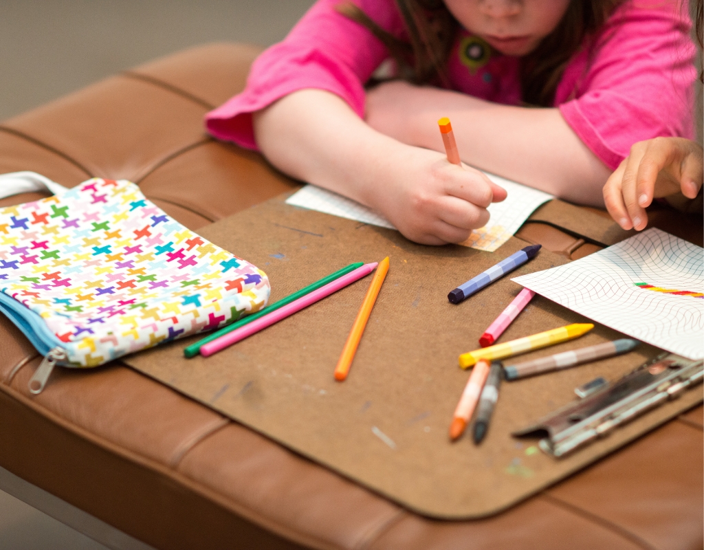 Close-up of a child's hands as they draw with colorful pencils on a leather bench. Their arms are visible in the trop of the frame.