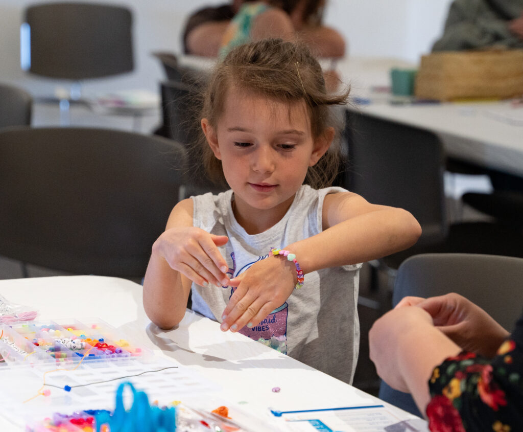 Photograph of a child making a bracelet with colorful beads on a white table. To the right of the frame are a pair of adult hands.