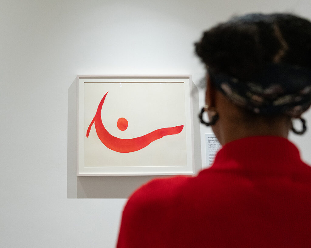 Photograph of a person with dark hair in buns wearing a bright red sweater from behind as they look at a painting by O’Keeffe on a white gallery wall. The painting shows a wave-like line with a dot above in shades of red.