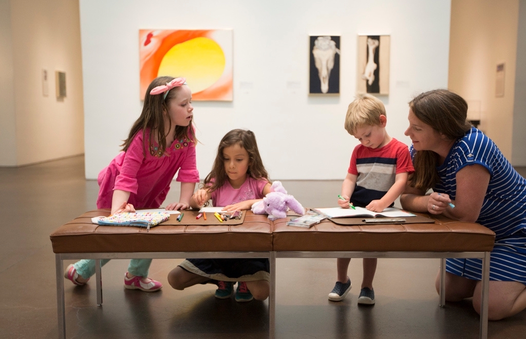 Photograph of three children standing or squatting at a bench in a gallery. The children are drawing on pieces of paper with colorful pencils. To the right is an adult crouched near them.