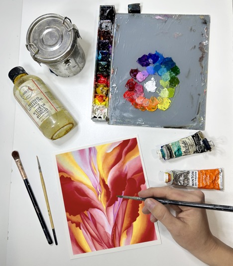 Photograph from above of a person's hands as they paint some abstracted florals in shades of red. Around their canvas are paints, brushes, oil, and a palette.