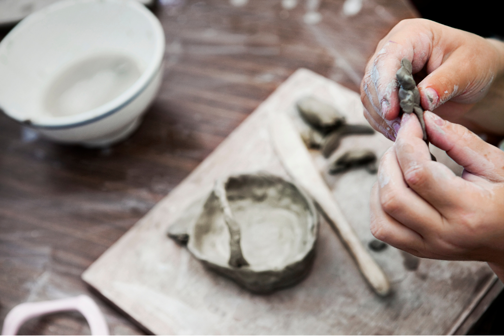 Photograph of hands shaping grey clay above a table where tools and clay are in place to make a bowl.