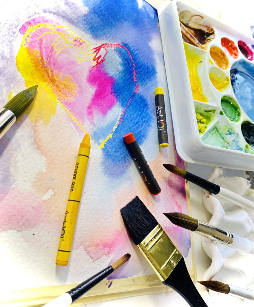 Photograph of a heart made with pastel along with watercolor shapes on a canvas. On top are brushes, pastels and a palette.