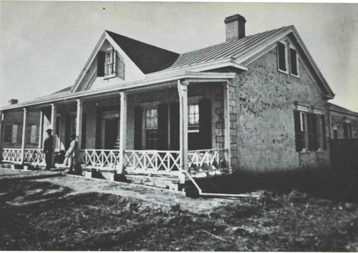 Black and white photograph of a house with a porch. Two figures stand in front.