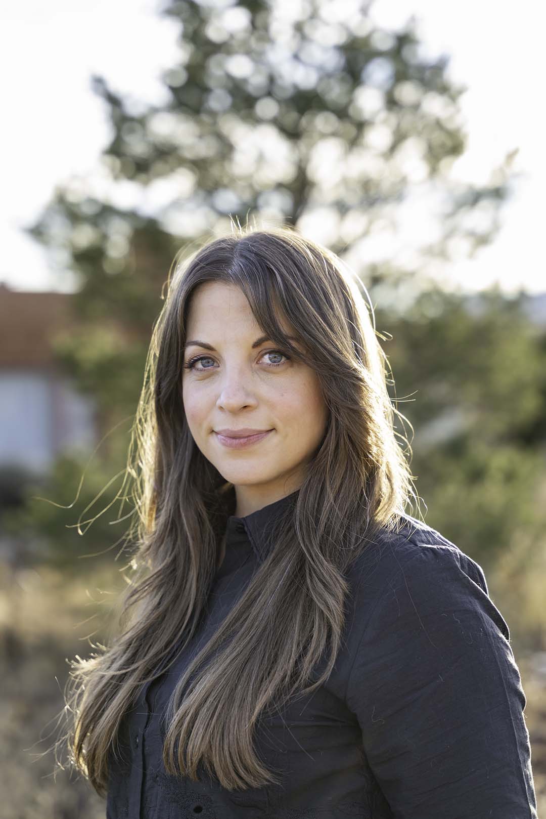 Photograph of a person with long brown hair and a dark jacket standing in front of a blurry adobe home and tree. They are smiling at the camera.