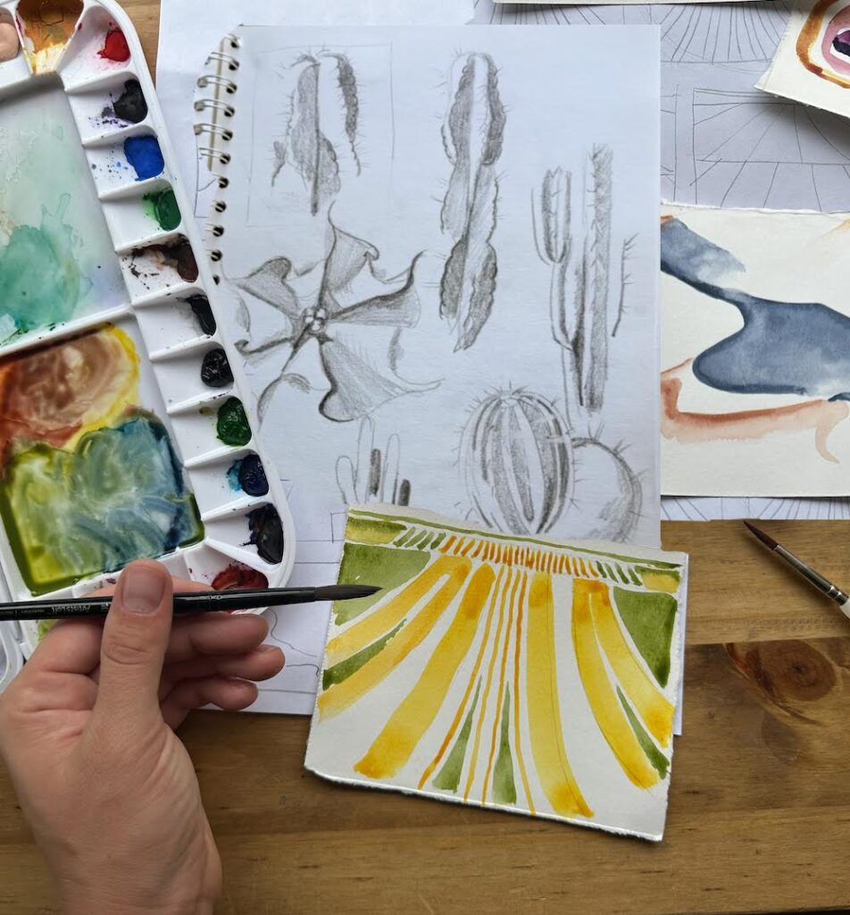 Photograph of an artist's hand as they paint a small canvas in shades of green and yellow. Below their canvas are charcoal sketches of cacti and to the right is a watercolor palette.