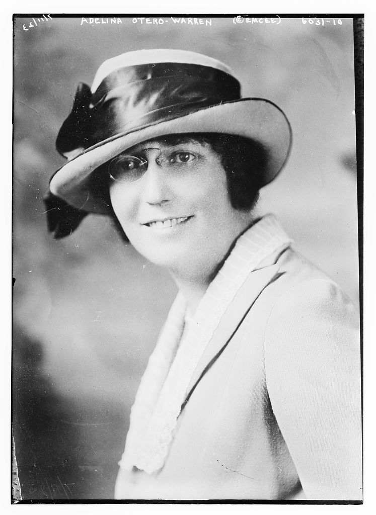 Black and white portrait of a person with small spectacles and a large hat with a ribbon smiling at the camera.