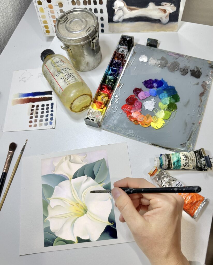 Photograph of an artist's hands as they paint a white flower on a small canvas. Above their hand is an oil pain palette, Lindseed oil, tubes of paint, and other artist supplies.