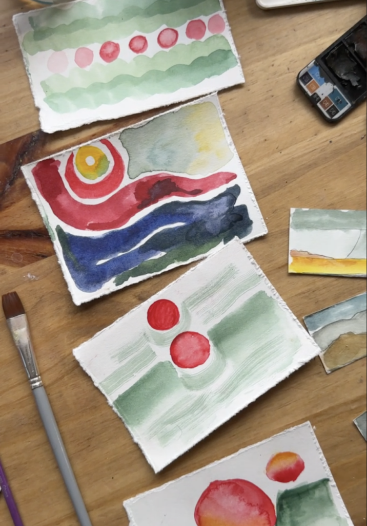 Photograph of three pieces of paper with watercolor paintings of abstract landscapes in shades of red, green, yellow and red. Near the papers are paintbrushes and art supplies. They all sit on a wooden table