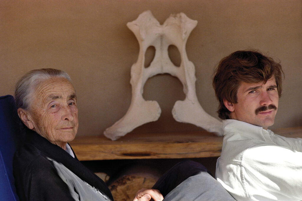 Georgia O'Keeffe with her grey hair tied up sitting in front of Juan Hamilton. Both sit in front of a large white pelvis bone propped on an adobe wall and both look in the direction of the camera.