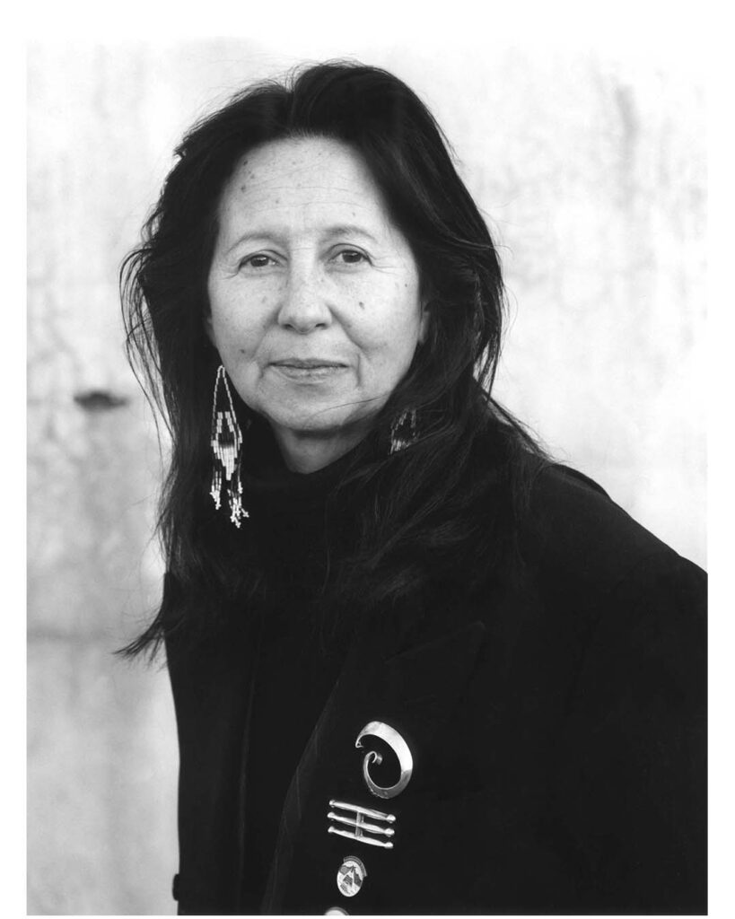 A black and white portrait of a person with long dark hair staring at the camera and smiling slightly. They wear a dark jacket with intricate brooches and long earrings. 