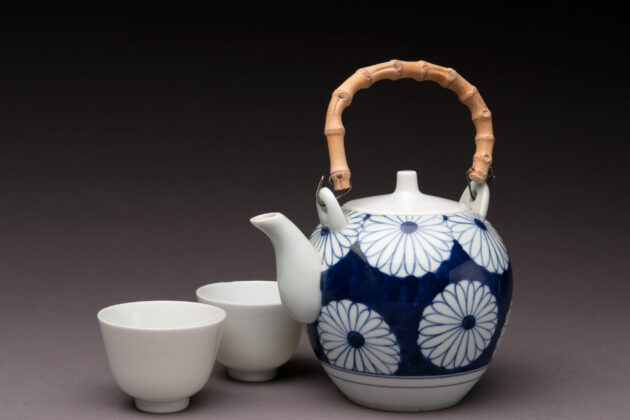 A photograph of a blue daisy floral pattern Japanese teapot with a bamboo handle. On the left are two simple ceramic teacups.