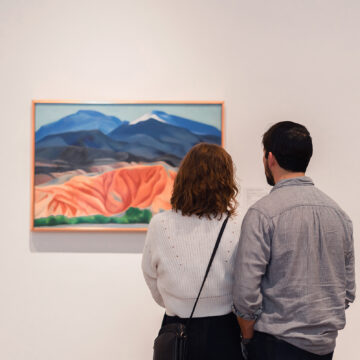 : A photograph of two people taken from behind, they are standing close together looking at O’Keeffe’s painting ‘Black Mesa Landscape, New Mexico / Out Back of Marie's II’ in the Museum galleries.