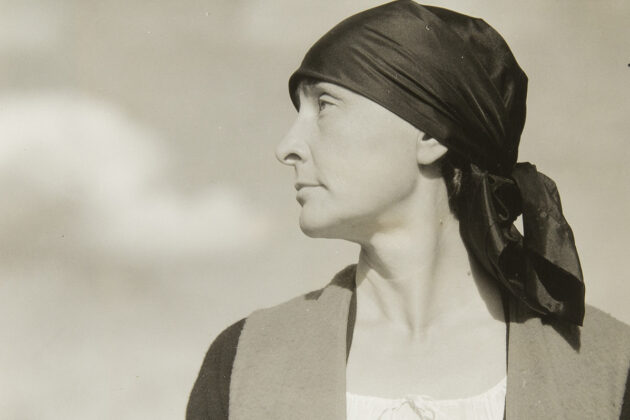 Half-length portrait of Georgia O'Keeffe with head in profile facing viewer left wearing a dark scarf/ turban tied behind her head; torso is frontal, neutral facial expression with eyes looking upward; she wears a white scoop-neck drawstring blouse under a dark sweater and vest; framed against a sky with clouds