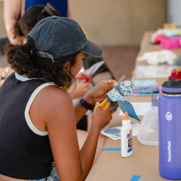 Young person participating in an arts and craft activity. They are wearing a hat and holding some paper and scissors.