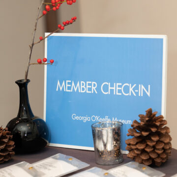 Photograph of a blue 'Member Check-In' sign on a table surrounded with candles, pinecones, and branches in a vase. On the bottom of the frame are name tags