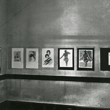 Black and white photograph showing several of O'Keeffe's watercolors on display. On the wall in focus are eight of O'Keeffe's watercolors, including a wall of an exhibition with three nude watercolors and several more. Under the hanging paintings is a dark moulding.