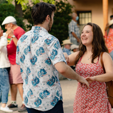 Photograph of two people dancing and smiling together holding hands. Behind them are trees, an adobe building and other couples dancing.