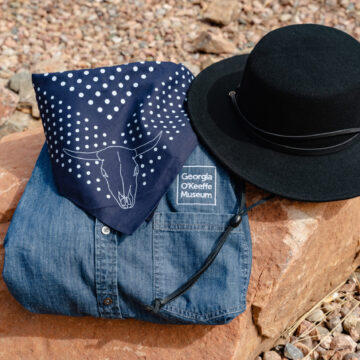 Photograph of a folded blue denim shirt with the 'Georgia O'Keeffe Museum' Loago on the breast pocket, a spotted blue bandana, and a black gaucho hat on a large red rock and surrounded by smaller stones.