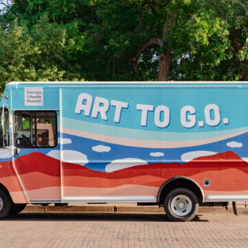 Photograph of a truck with a desert horizon and cloud motifs on the side along with the heading 'Art to G.O.' and the Georgia O'Keeffe Museum Logo. Behind the truck are crowd of people under green trees.