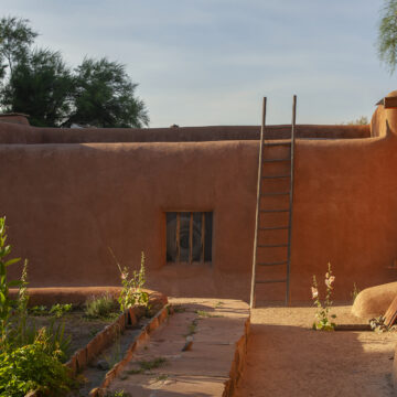 Photograph of O'Keeffe's home in Abiquiú where an adobe wall with a window in the center sits behind a raised green garden. On the wall leans a wooden ladder. Behind the adobe wall a tree and blue sky is visible.