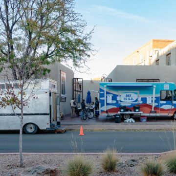 Photograph of a street and an adobe building with a tree on the left of the frame. Parked in front of the building and on the street are two trucks, one a grey mobile art gallery and one the Art to G.O. truck with cloud motifs on it and two tables in front.
