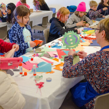 Photograph of a long table with several children and a few adults making art pieces with colorful paper.