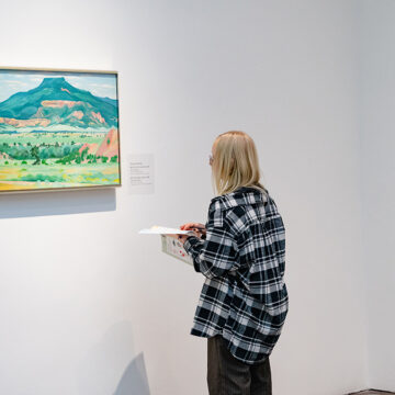 A young person with long hair and glasses holding a paper and pen and looking at a painting on a white gallery wall. The oil painting by O’Keeffe is of a flat-topped mountain in fall, predominantly in dark greens and oranges.