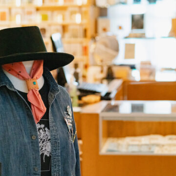 A mannequin with a black gaucho hat, a red tied scarf, a t-shirt, and a denim vest. Behind the mannequin in the background are shelves of the Museum Store.