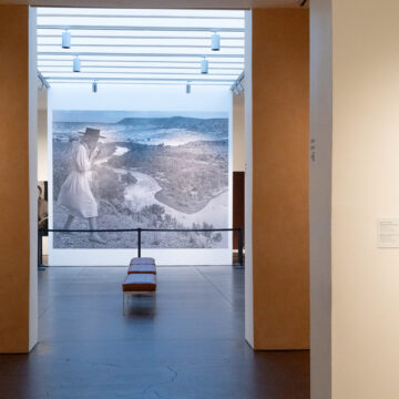 Interior of the Museum with a bench in the room farthest from the camera. In the farthest gallery is a large mural with a black and white photograph of O'Keeffe. On the nearest wall to the right is a small painting of a pink flower by O'Keeffe.