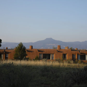 Photograph of the exterior of an adobe home with several windows. Behind the home is a mountain across the horizon and a clear blue sky.