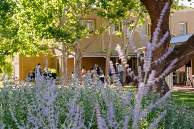 Photograph of a group of people on the patio of an adobe building. The people are in the background and blurry. They are framed by green trees and purple lavender plants in the foreground.