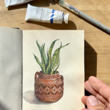 Photograph of a small book open to a page with a watercolor of a pot with a plant in it. To the right of the page a hand holds the book open. Above the book are small paint tubes and a paintbrush.