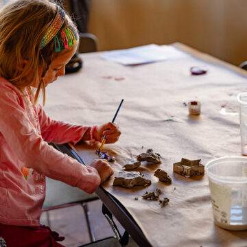 Photograph of a child sitting at a table making art with pieces of clay and a paintbrush.