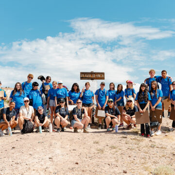 A group of young people stand or squat in two rows in front of a wooden sign that highlights the 'Plaza Blanca' or 'White Place.' Many of them wear hats and sunglasses. Many of then wear blue t-shirts marking them as.a cohesive group. Their environment is a dry desert landscape and behind the group is a blue sky with a few white clouds.