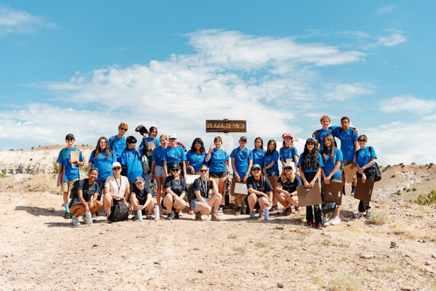 A group of young people stand or squat in two rows in front of a wooden sign that highlights the 'Plaza Blanca' or 'White Place.' Many of them wear hats and sunglasses. Many of then wear blue t-shirts marking them as.a cohesive group. Their environment is a dry desert landscape and behind the group is a blue sky with a few white clouds.