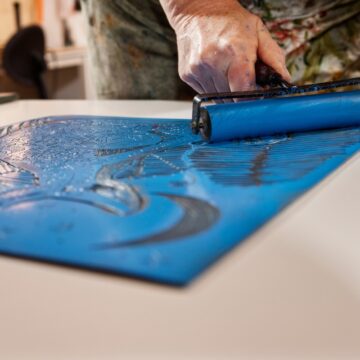 Photograph of a person's hand holding a brush as they spread blue ink on a carved material and prepare to make a print.