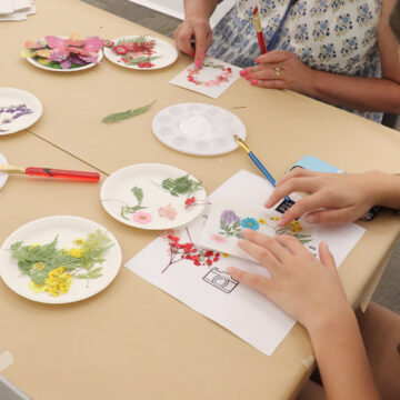 Photograph of a young person's hands as they glue flowers to a piece of paper on a table. To their right another person is pressing a brush against a piece of paper.