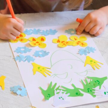 Close-up photograph of a young person's hands as they draw on a piece of paper covered with colorful cut-outs