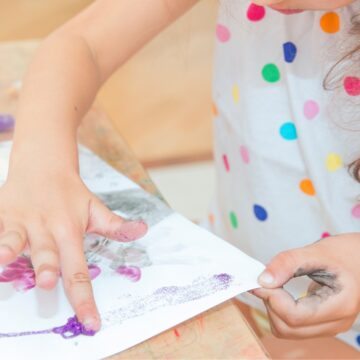 Photograph of a young child's hands as they use their fingers to paint a painting on a while piece of paper.